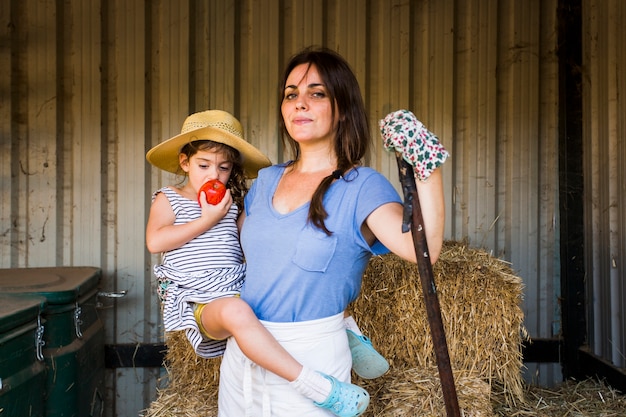 Foto gratuita madre llevando a su hija comiendo manzana roja de pie frente a la pila de heno