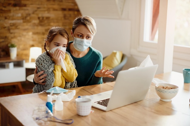 Madre con mascarilla y su hija enferma haciendo videollamadas con un médico desde casa