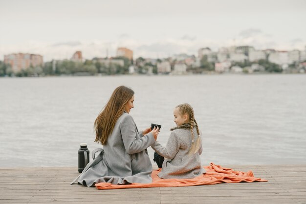 Madre de moda con hija. Gente de picnic. Mujer con un abrigo gris. Familia junto al agua.