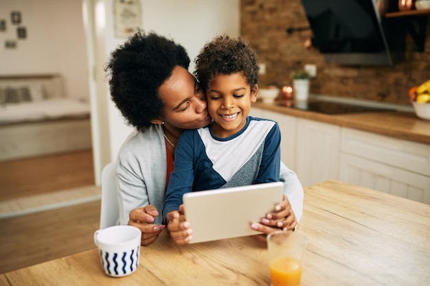 Madre negra afectuosa besando a su hijo mientras usa una tableta digital en casa