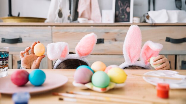 Madre con niño en orejas de conejo escondido detrás de una mesa con huevos de colores