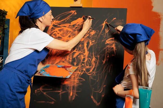 Madre con un niño pequeño creando una obra maestra con pintura aquarelle de color naranja y paleta de tintes con pincel. Niña pequeña aprendiendo habilidades de pintura sobre lienzo usando acuarela e inspiración artística.