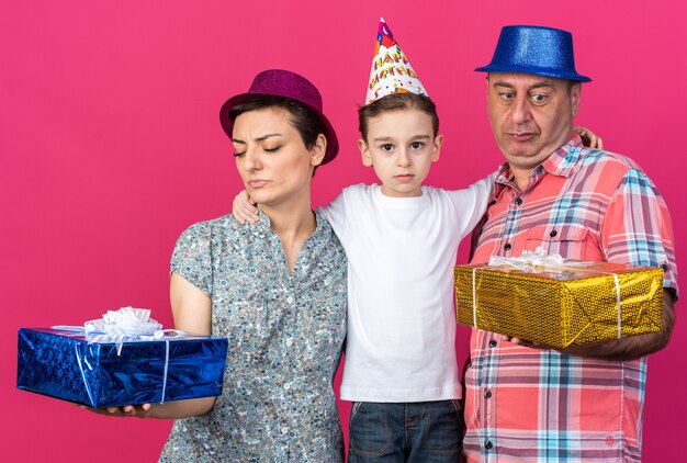 Madre y padre descontentos con sombreros de fiesta sosteniendo y mirando cajas de regalo de pie con su hijo aislado en la pared rosa con espacio de copia