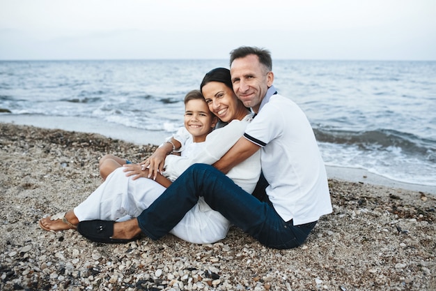 Madre, padre e hijo están sentados en la playa cerca del mar, abrazados y mirando directamente