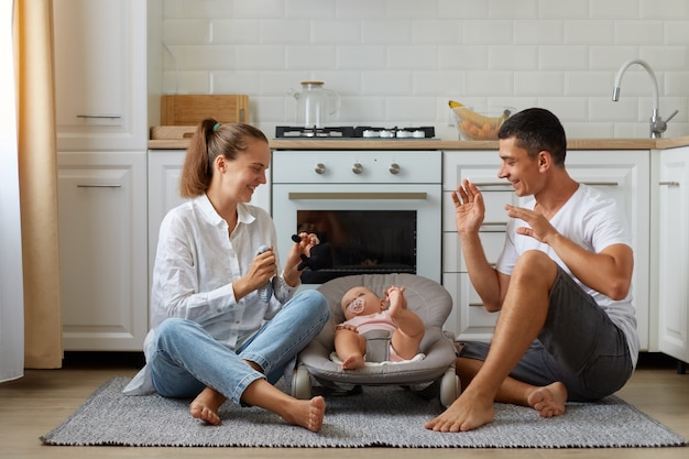 Madre y padre jugando con su hijo o hija en una mecedora en el piso de la sala de luz con cocina en el fondo, familia feliz pasando tiempo juntos, jugando con el bebé.