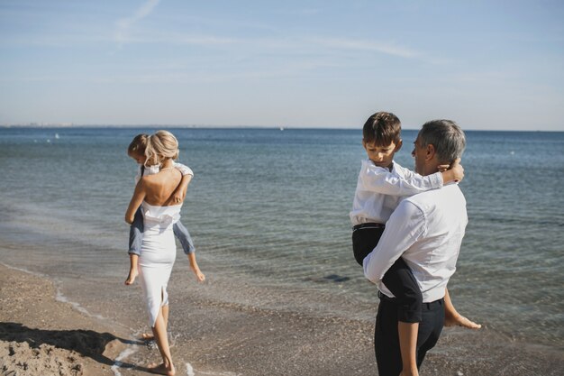 Madre y padre sostienen a sus hijos pequeños en las manos en el soleado día de verano, caminando junto al mar