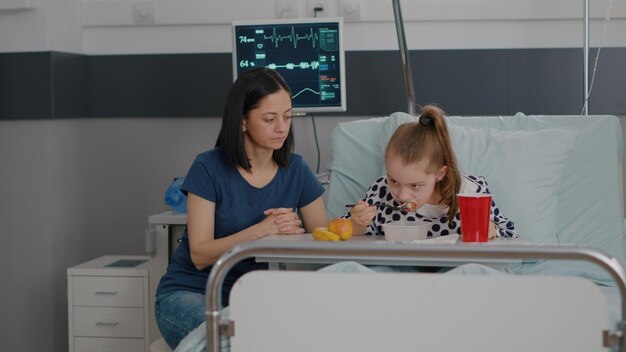 Madre de pie con su hija enferma mientras come comida sana durante el almuerzo durante el examen en la sala del hospital. Niña hospitalizada recuperándose después de una cirugía médica con desayuno nutricional