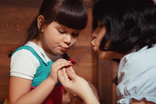 Madre pintando las uñas de su hija. Closeup retrato de madre e hija haciendo maquillaje aislado sobre fondo de madera.