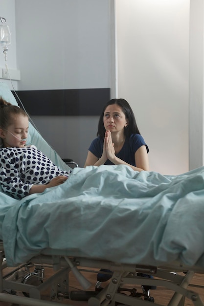 Madre preocupada rezando por una niña enferma descansando en la cama del paciente. Niño enfermo bajo tratamiento con medicamentos durmiendo dentro de la sala de pediatría de la clínica de salud mientras los padres esperan que ella se despierte.