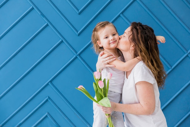 Madre que besa a su pequeña hija bonita que sostiene las flores del tulipán sobre fondo azul