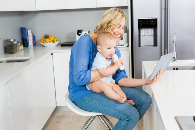 Madre que celebra al bebé durante el uso de la tableta digital en la cocina