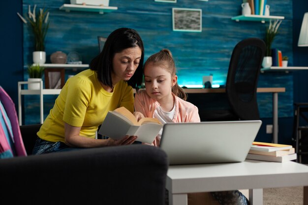 Madre sentada junto a su hija sosteniendo el libro de la escuela leyendo juntos