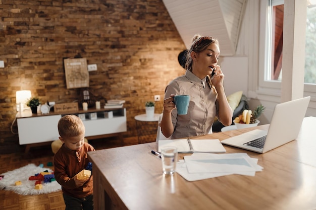 Madre soltera hablando por teléfono mientras usa la computadora y está con su hijo en casa