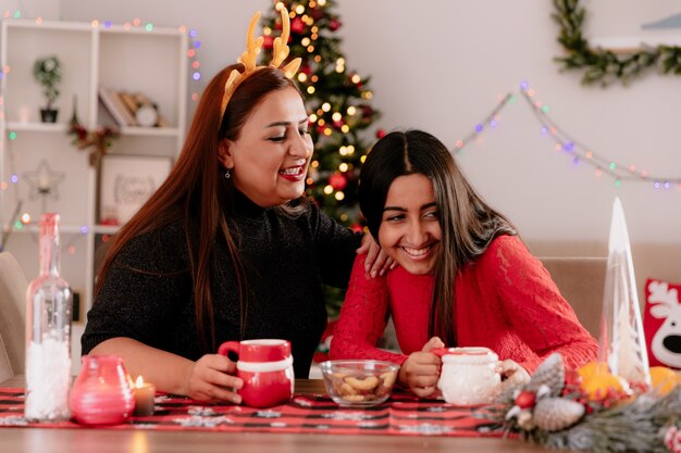 Madre sonriente con diadema de renos mira a su hija sentada a la mesa disfrutando del tiempo de Navidad en casa