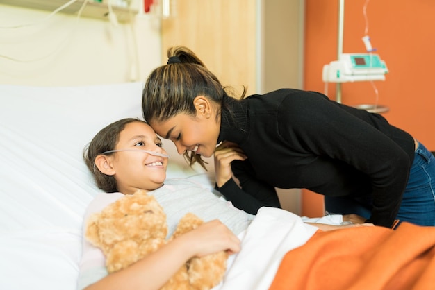Madre sonriente e hija enferma junto a la cama en el hospital durante la visita