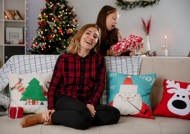 Madre sonriente mira de lado sentado en el sofá e hija emocionada sosteniendo el paquete de regalo y disfrutando de la Navidad en casa