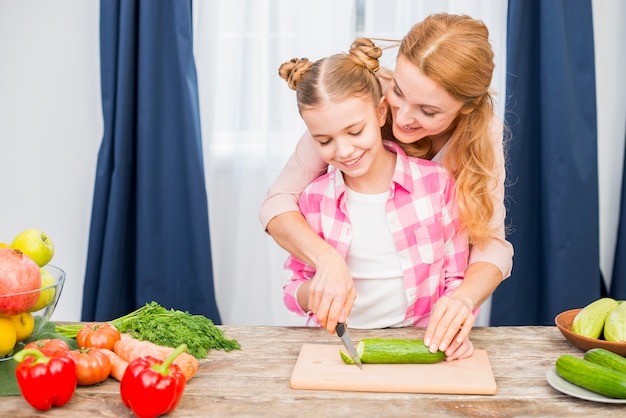 Madre sonriente que ayuda a su hija para cortar el pepino en la tabla de cortar