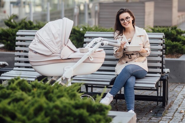 Madre sonriente sosteniendo una lonchera de plástico mientras está sentado en un banco con un cochecito y un bebé recién nacido