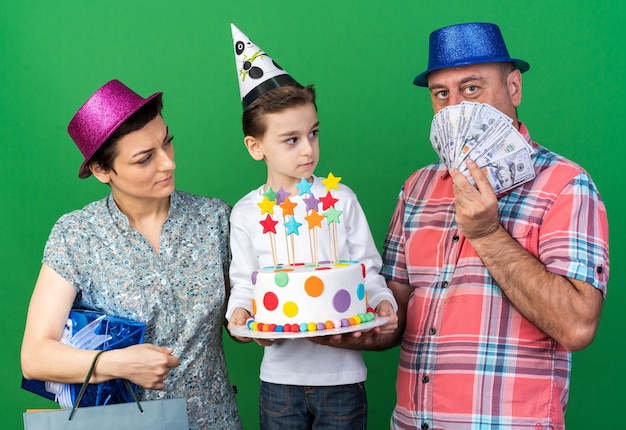Foto gratuita madre sorprendida con sombrero de fiesta púrpura con caja de regalo y su hijo con gorro de fiesta sosteniendo pastel de cumpleaños mirando al padre con sombrero de fiesta azul y sosteniendo dinero en la pared verde