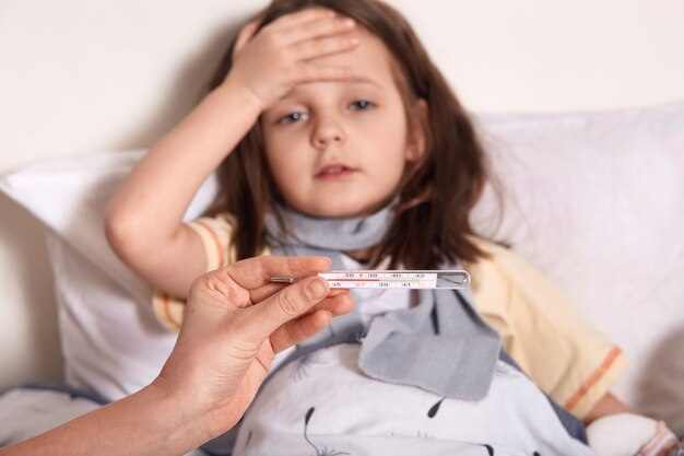 Madre sosteniendo el termómetro en las manos, niña enferma acostada en la cama y manteniendo la palma de la mano en forehea, sufriendo de alta temperatura