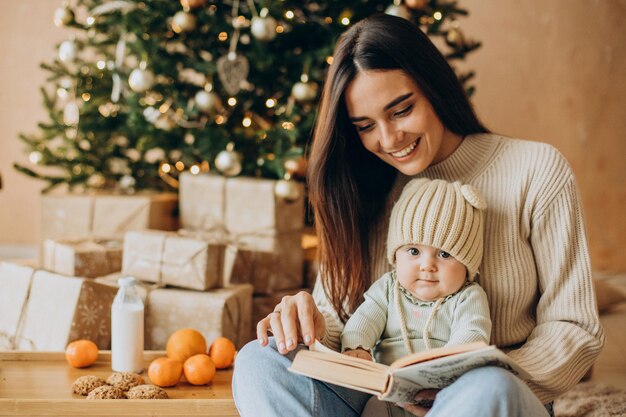 Madre con su hija leyendo un libro junto al árbol de Navidad