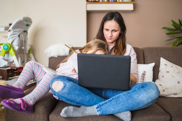 Madre con su hija sentada en el sofá usando la computadora portátil