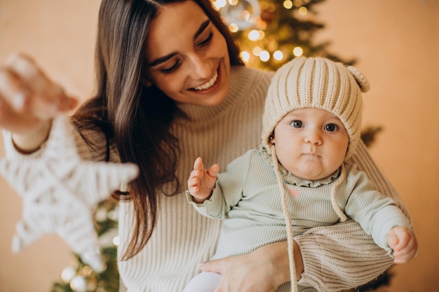 Madre con su hija sosteniendo juguetes navideños junto al árbol de navidad