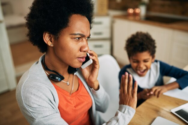 Madre trabajadora afroamericana disgustada hablando por teléfono móvil en casa