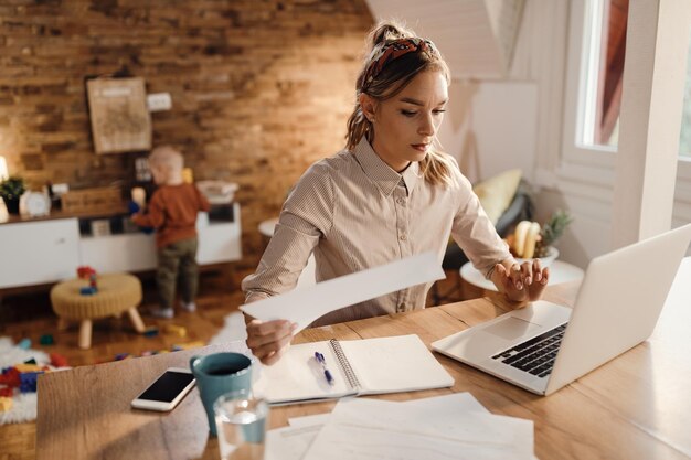 Madre trabajadora que usa una computadora portátil mientras revisa informes comerciales en casa