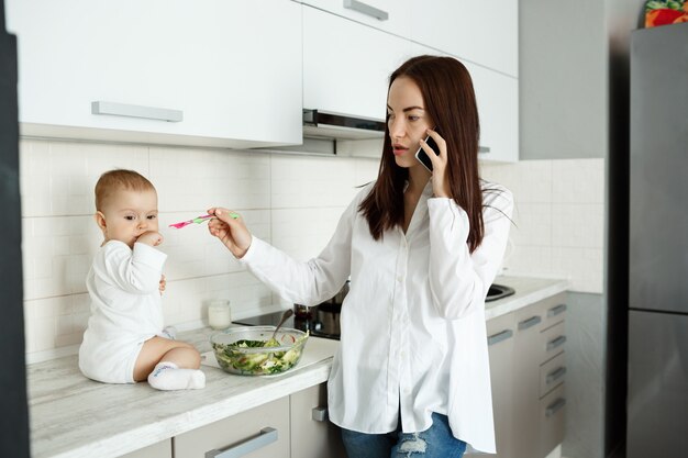 Madre trabajando desde casa, hablando por teléfono y alimentando a un lindo bebé