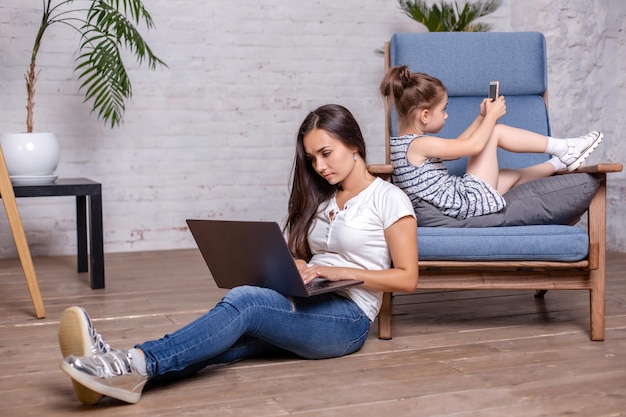 Madre trabajando con una laptop, sentada en el suelo y una linda niñita tendida en el sillón y jugando juegos en el smartphone
