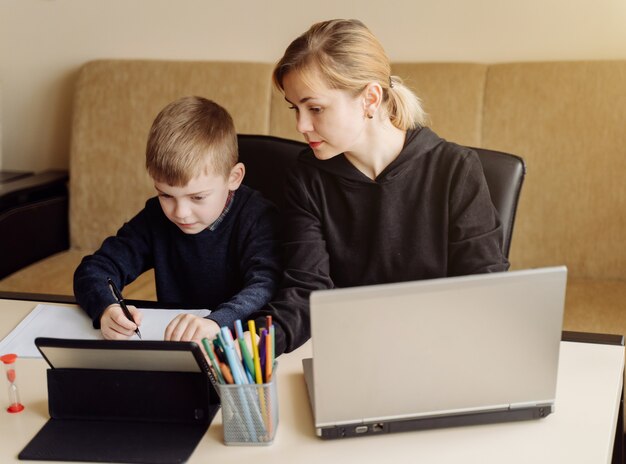 Madre usando una computadora portátil y una tableta enseñando con su hijo en línea en su casa en su habitación