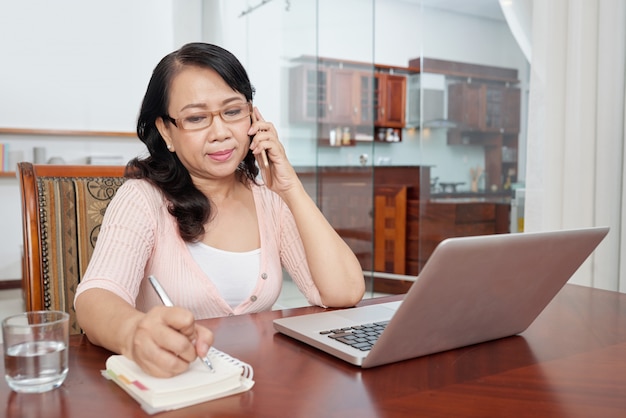 Madura mujer asiática sentada en la mesa en casa con el portátil, hablando por teléfono y tomando notas