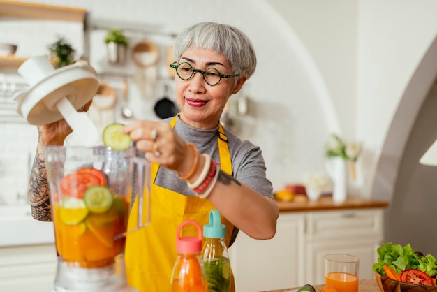 Madura mujer tatuada sonriente comiendo ensalada de frutas y verduras Atractiva mujer madura con ensalada de frutas verdes frescas en casa Delantal de mujer mayor de pie en el mostrador de la cocina relajándose en casa