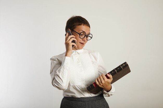 Maestra en blusa blanca y falda gris mira el gran libro que sostiene mientras habla por su teléfono móvil aislado en blanco