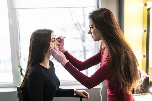 Maestra haciendo maquillaje profesional de cejas de mujer joven