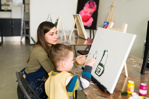 Una maestra hispana le está dando instrucciones a un niño pequeño caucásico para que use un pincel y haga una pintura en un lienzo en blanco para una clase de arte