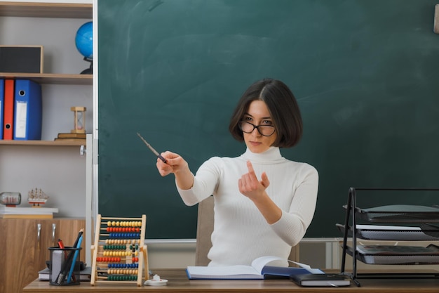 Una maestra joven y confiada que usa anteojos apunta a un lado con un puntero sentado en el escritorio con herramientas escolares en el aula