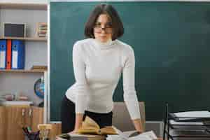 Foto gratuita una maestra joven y estricta con anteojos parada frente a la pizarra en el aula