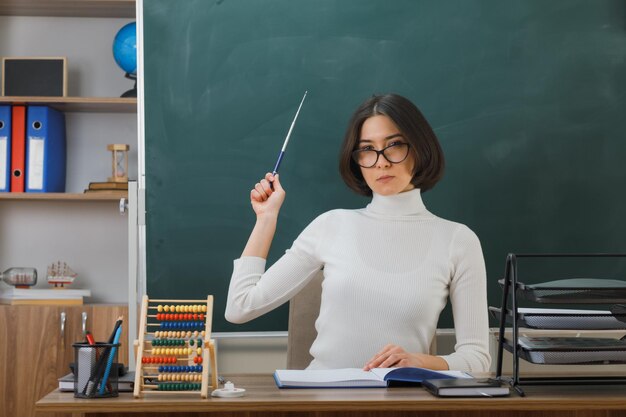 Una maestra joven y estricta que usa gafas apunta con un puntero en la pizarra sentada en el escritorio con herramientas escolares en el aula