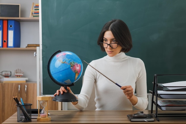 una maestra joven y estricta señala el globo terráqueo con un puntero sentado en el escritorio con herramientas escolares en el aula