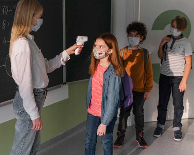 Maestra con máscara médica que controla la temperatura de los niños en la escuela