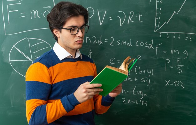 Maestro de geometría joven concentrado con gafas de pie delante de la pizarra en el libro de lectura del aula