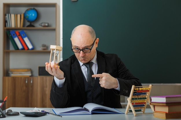 maestro hombre con anteojos sentado en el escritorio de la escuela frente a la pizarra en el aula sosteniendo un reloj de arena explicando la lección mirando con el ceño fruncido