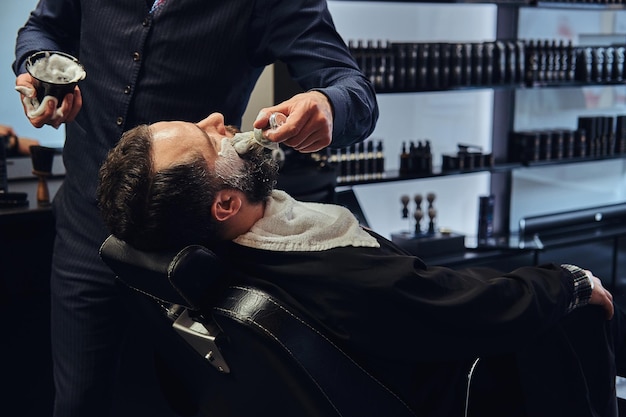 Un maestro peluquero prepara la cara para afeitarse y unta la cara con espuma en un salón de peluquería.