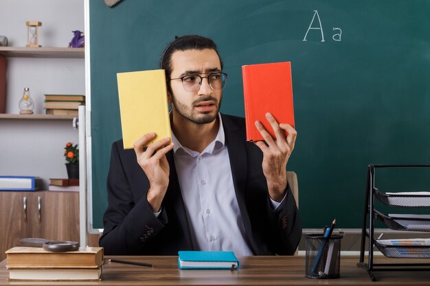 Maestro preocupado con gafas sosteniendo y mirando el libro sentado a la mesa con herramientas escolares en el aula