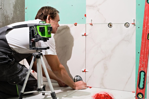 Maestro profesional colocando baldosas de cerámica en la pared del baño Retrato de un reparador experimentado colocando baldosas de porcelana de gran tamaño Trabajador de la construcción comprobando la instalación de baldosas con nivel láser