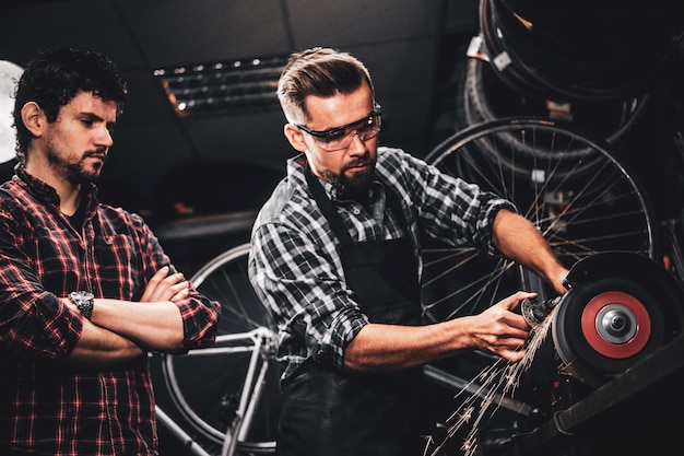 El maestro de reparación experto está trabajando con una máquina herramienta en un taller de bicicletas ocupado y su colega está parado cerca.