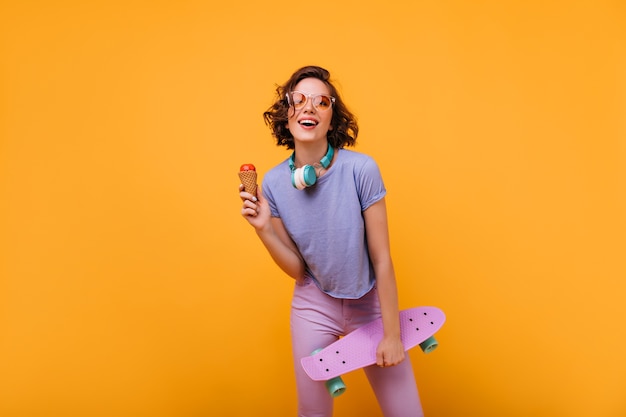 Magnífica chica blanca en ropa casual posando en auriculares. Retrato de bella dama con patineta comiendo helado.