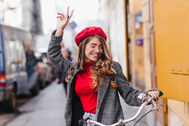 Magnífica chica elegante en suéter rojo que expresa verdaderas emociones sentado en bicicleta
